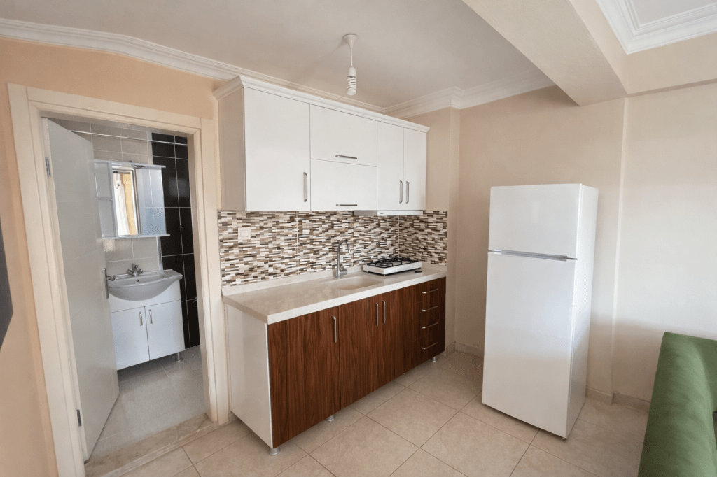 Modern kitchen with white and brown cabinets, a gas stove, and a refrigerator, featuring a tiled backsplash, and a doorway leading to a bathroom.