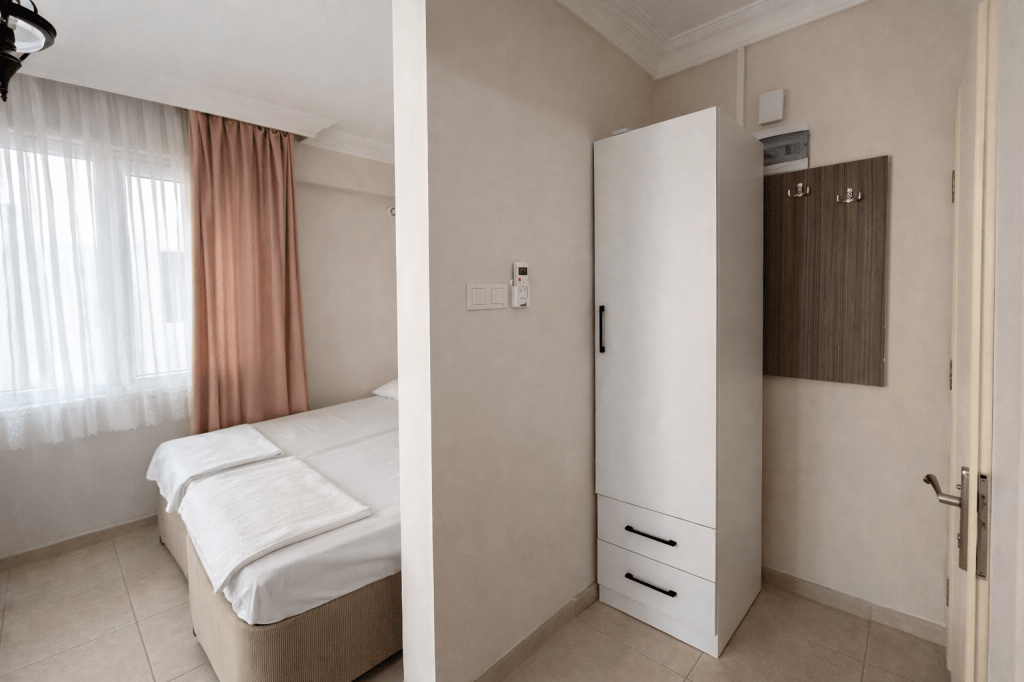 A small, modern bedroom featuring a bed with white linens, a large window with sheer curtains, a white wardrobe, and a wooden wall panel with hooks.