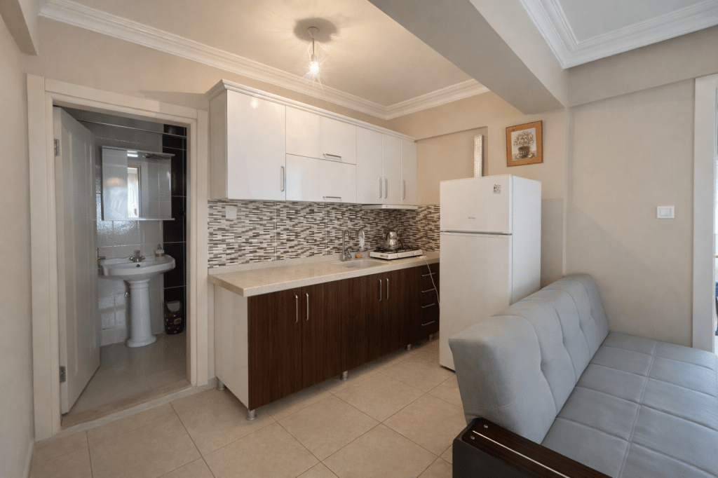 Modern kitchen with white cabinets, a gray sofa, and a small bathroom visible in the background.