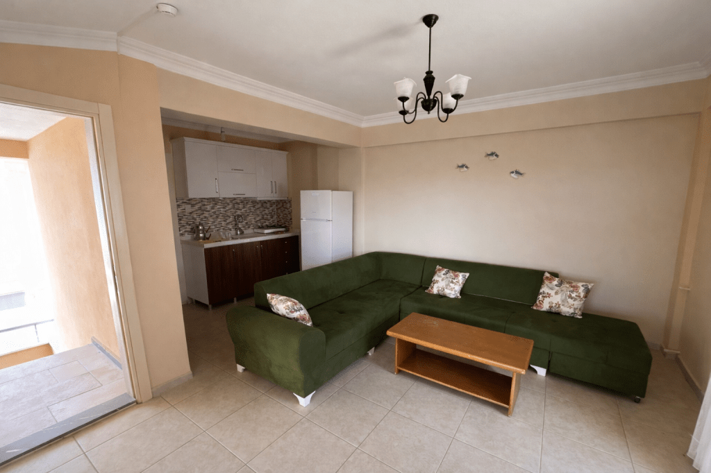 A cozy living room featuring a green sectional sofa with floral cushions, a wooden coffee table, and a light fixture, with a view into an adjoining kitchen area.