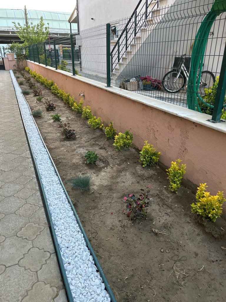 A landscaped garden area with a row of small shrubs and flowers, bordered by a white pebble pathway, alongside a building with a staircase and a bike parked in the background.