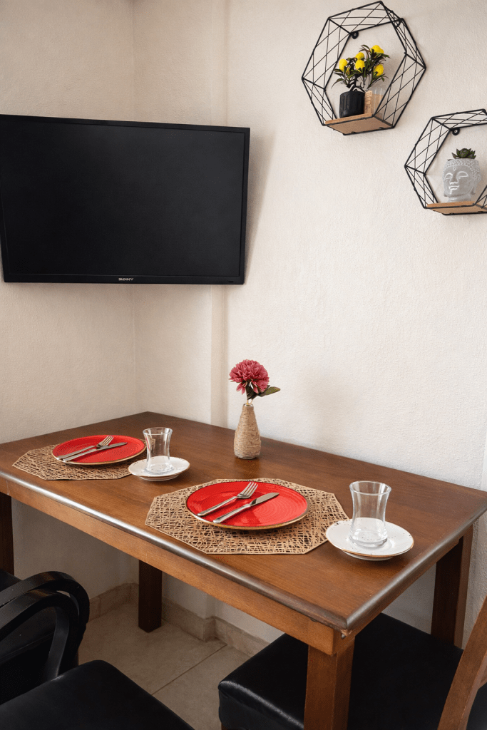 Cozy dining area featuring a wooden table set for two with red plates, cutlery, and glassware, complemented by a decorative flower vase and wall-mounted plant holders.