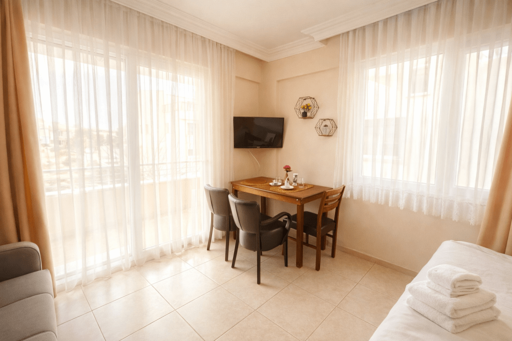 A cozy hotel room featuring a small dining table with two chairs, a television mounted on the wall, large windows covered by sheer curtains, and a comfortable sofa. The decor is light and airy, creating a welcoming atmosphere.