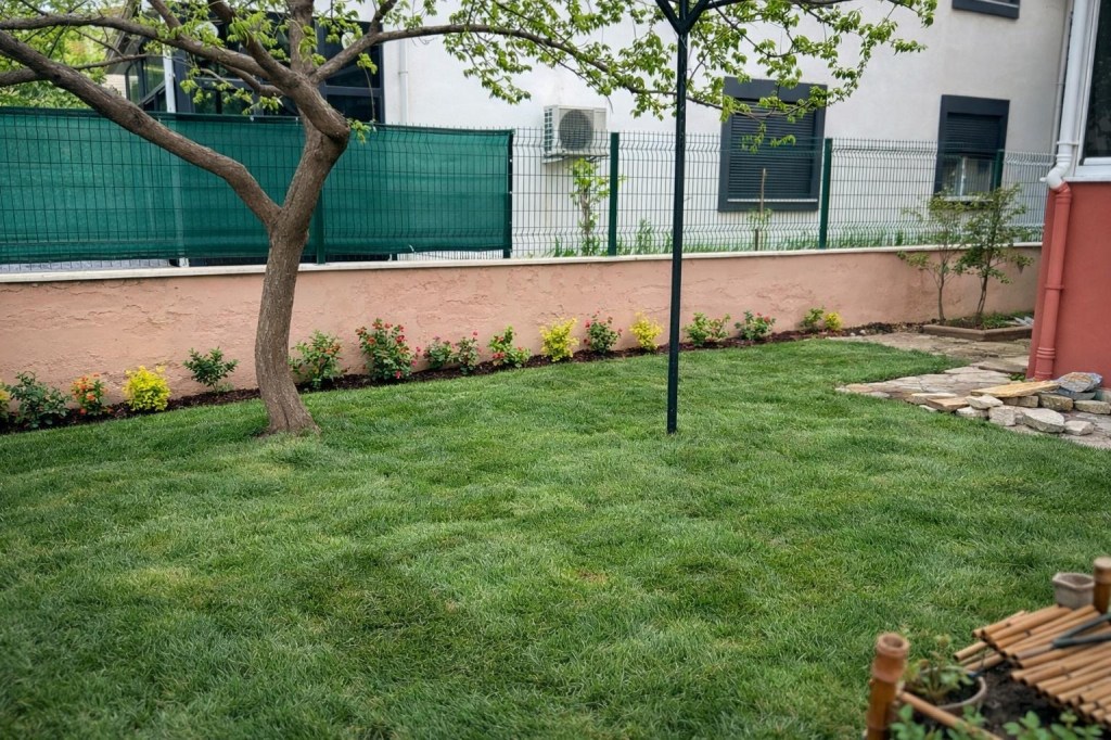 A well-maintained garden with lush green grass, a small tree, and a flower border along a pink wall. There are some decorative stones and garden tools in the foreground.
