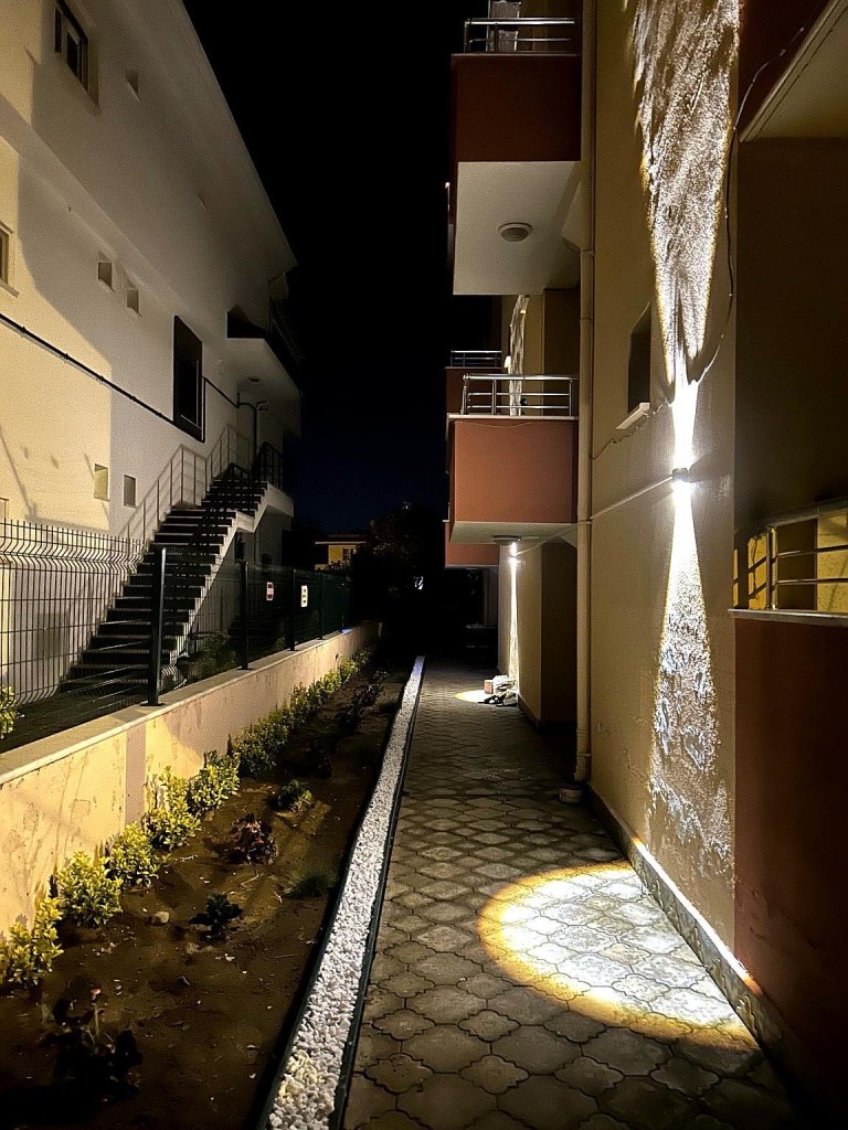 A narrow illuminated pathway between two buildings at night, featuring a landscaped area with plants and a light casting shadows on the walls.