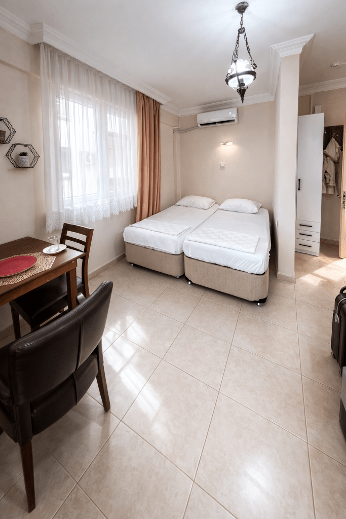 Interior view of a small room featuring two single beds, a dining table with a chair, and a suitcase, with a window covered by sheer curtains.