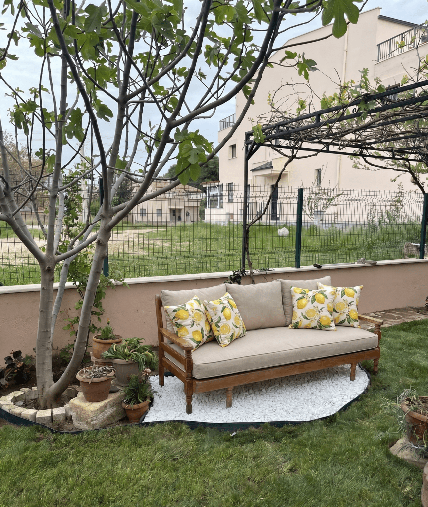 A comfortable wooden outdoor sofa with lemon-patterned pillows, surrounded by vibrant potted plants in a grassy garden setting, with a house and fence in the background.