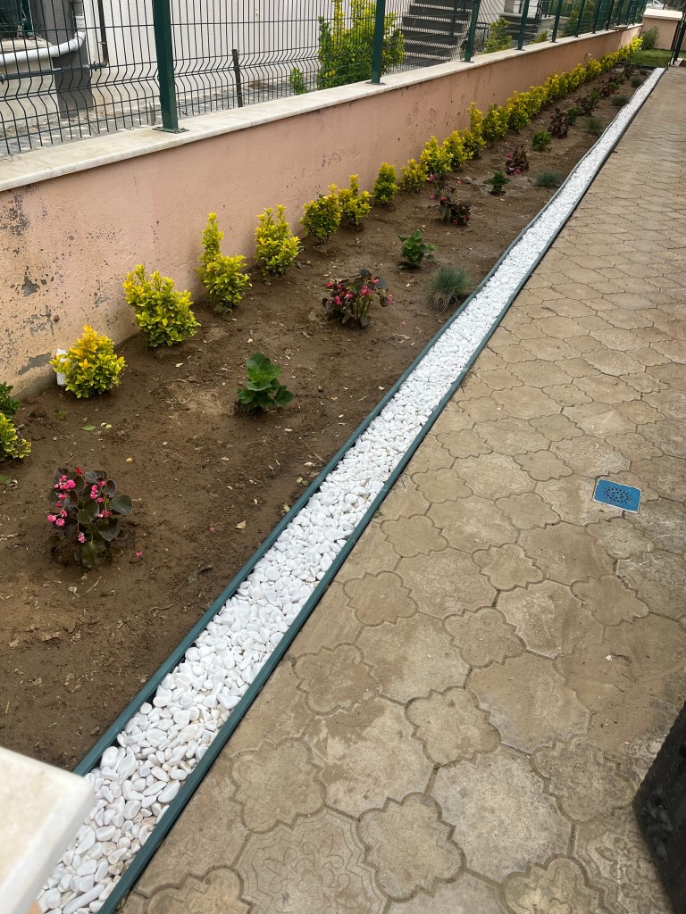 A landscaped garden featuring a row of small flowers and plants along a path, bordered by white pebbles and a pink wall.